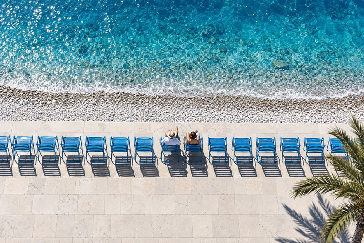 Love room à Nice : couple assis sur les chaises bleues de la promenade des Anglais face à la mer turquoise