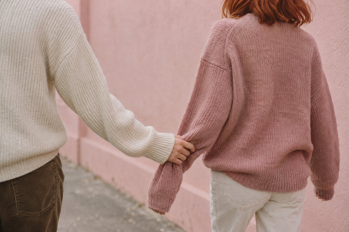 Style d'attachement : une main retient la manche du pull de l'autre, deux silhouettes de dos devant un mur rose poudré